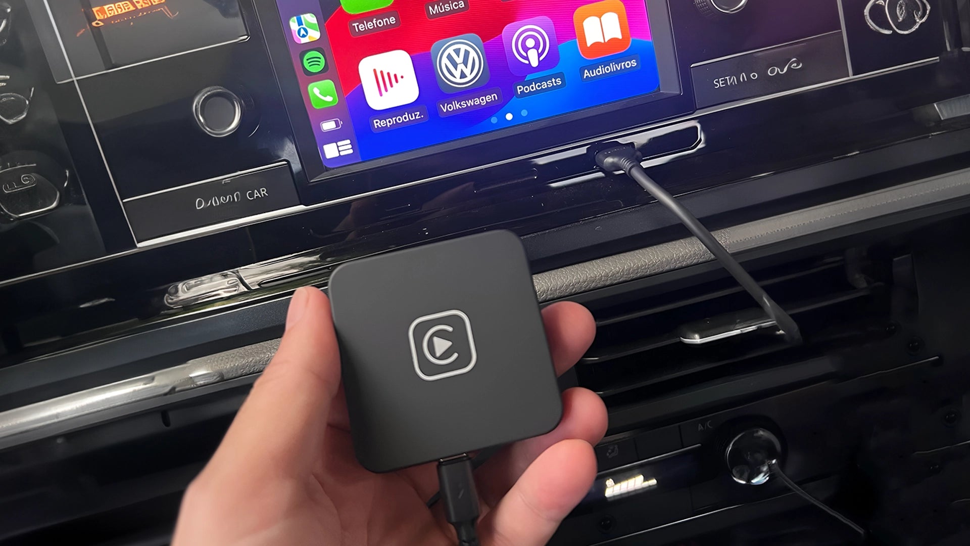 Hand holding a CarPlay adapter in front of a car's multimedia system with the CarPlay interface visible on the screen, indicating the adapter's compatibility and use.
